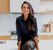 A woman smiling while sitting on a counter, wearing casual clothing, with focus on her face and neck area, suggesting a relaxed, nurturing atmosphere.